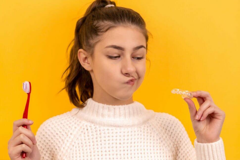 Brunette girl who holds a toothbrush and clear orthodontic aligners against a yellow background