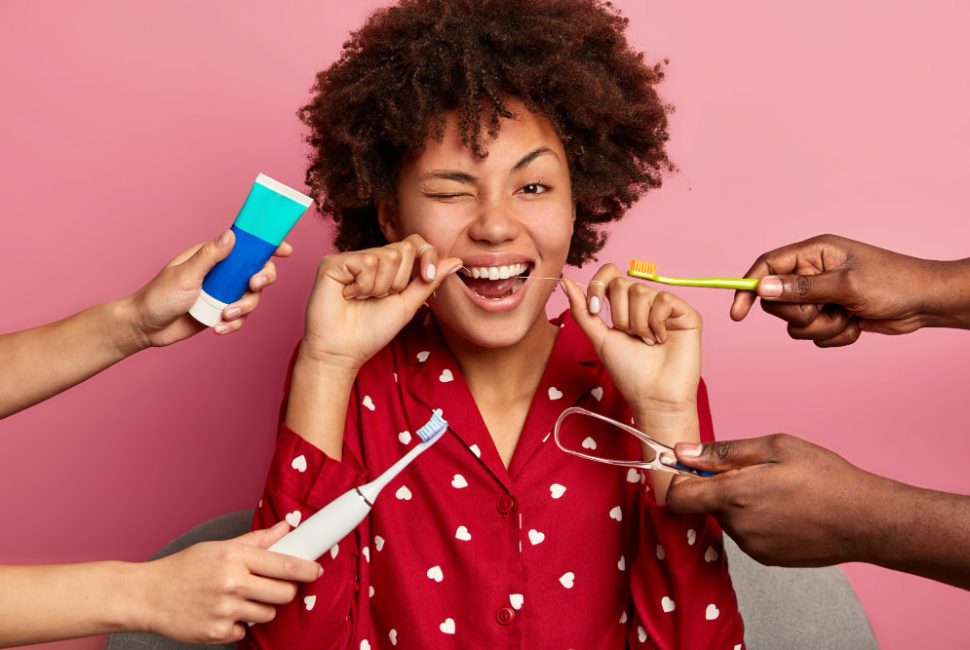 Young woman using tooth floss surrounded with toothpaste, electric toothbrush and tongue cleaner